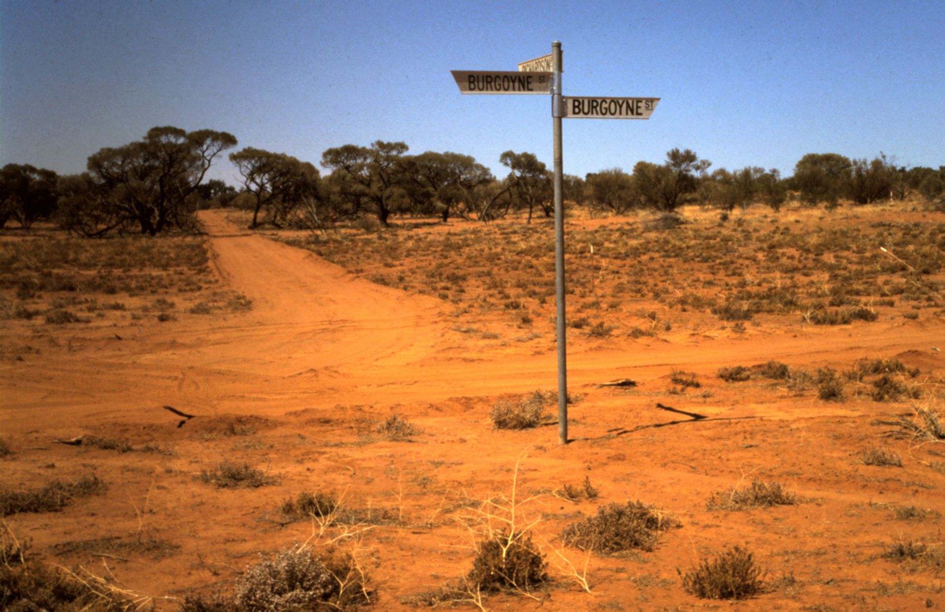 A historical photo showing the original location of Richardson Place, now the main street in Roxby Downs, viewed from what is currently the hospital site. The image features a barren, red desert landscape with sparse vegetation, a dirt path, and a street sign reading 'Burgoyne St.' against a clear blue sky
